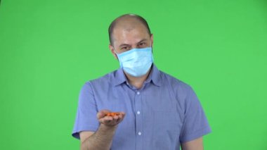 Medikal maskeli orta yaşlı bir adamın portresi kameraya ilaç taşıyan bir eli gösteriyor. Mavi gömlekli kel adam stüdyoda yeşil ekranda poz veriyor. Kapat..