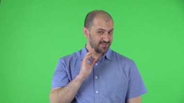 Fotoğrafta orta yaşlı adamlar kameraya bakıp el sallıyor ve buraya gelip göz kırpıyor. Mavi gömlekli kel erkek stüdyoda yeşil ekranda poz veriyor. Kapat..