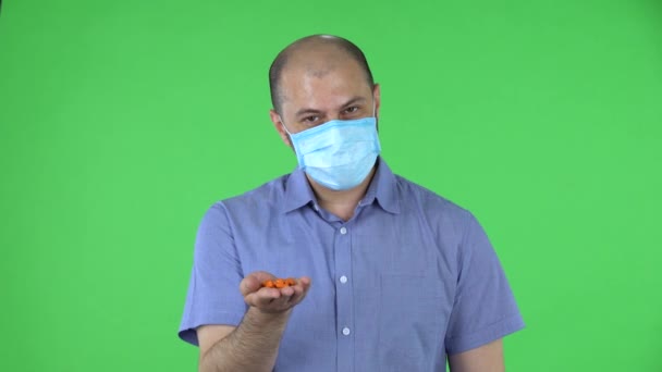Portretul unui bărbat de vârstă mijlocie în mască medicală arată o mână cu medicamente pentru cameră. Bărbat chel în cămașă albastră pozând pe ecran verde în studio. Close up . — Videoclip de stoc