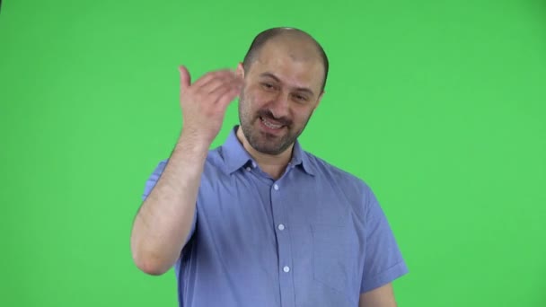 カメラを楽しそうに見ている中年男性の肖像画は、彼の手を振って、ここに来るためのジェスチャーを示しています。スタジオ内の緑のスクリーンにポーズをとった青いシャツに髭を生やした男。閉じろ!. — ストック動画