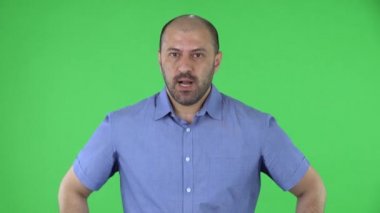 Orta yaşlı bir adamın portresi kameraya bakıyor ve sonra çok üzgün. Mavi gömlekli kel erkek stüdyoda yeşil ekranda poz veriyor. Kapat..