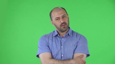 Kameraya bakan orta yaşlı bir adamın portresi çapraz kolların hayır anlamına geldiğini gösteriyor. Mavi gömlekli kel erkek stüdyoda yeşil ekranda poz veriyor. Kapat..