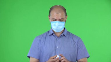 Medikal maskeli orta yaşlı bir adamın portresi ellerine dezenfektan veriyor. Mavi gömlekli kel adam stüdyoda yeşil ekranda poz veriyor. Kapat..