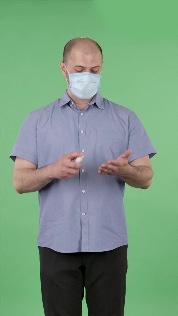 El retrato de un hombre de mediana edad con máscara médica trata sus manos con desinfectante. Hombre calvo con camisa azul posando en pantalla verde en el estudio. En cámara lenta. Vídeo vertical. — Vídeo de stock