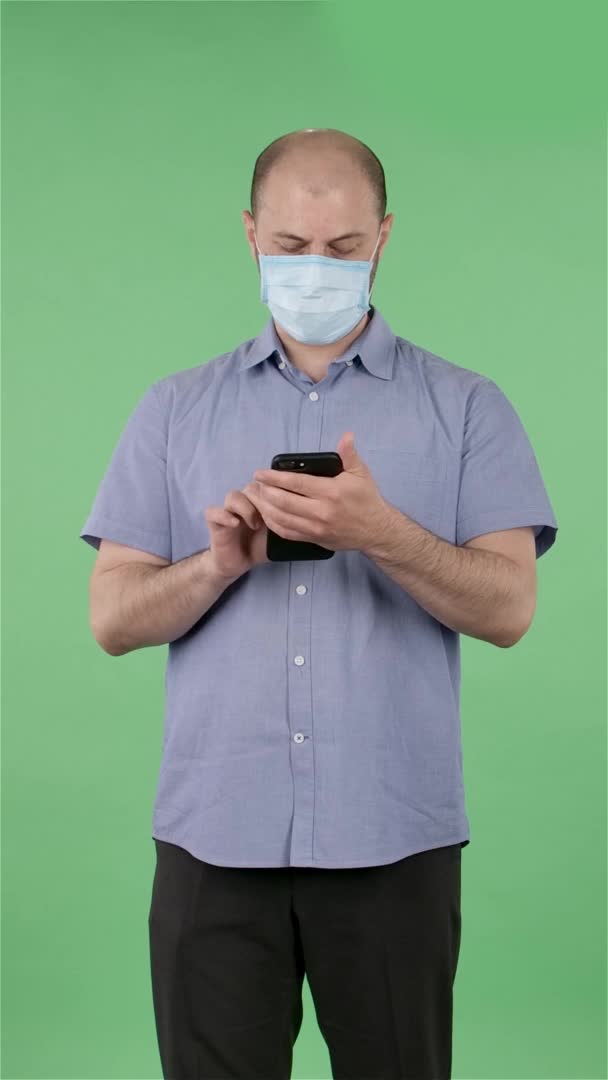 Ritratto di un uomo di mezza età in maschera medica che gli scrive sul telefono. Maschio calvo in camicia blu in posa sullo schermo verde in studio. Al rallentatore. Video verticale. — Video Stock