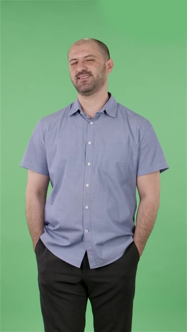 Portrait d'un homme d'âge moyen regardant la caméra flirte sourit ludique et clin d'oeil. Homme chauve avec une barbe dans une chemise bleue posant sur un écran vert dans le studio. Au ralenti. Vidéo verticale. — Video
