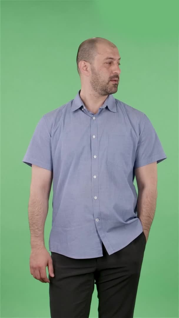 Portrait d'un homme d'âge moyen regardant autour de lui et faisant un geste silencieux secret. Homme chauve avec barbe en chemise bleue posant sur écran vert en studio. Au ralenti. Vidéo verticale. — Video
