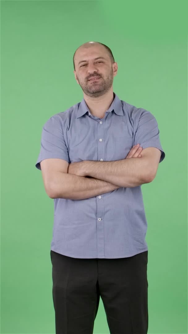 Портрет мужчины средних лет, смотрящего в камеру, весело улыбается и подмигивает. Лысый мужчина с бородой в синей рубашке позирует на зеленом экране в студии. Медленное движение. Вертикальное видео. — стоковое видео
