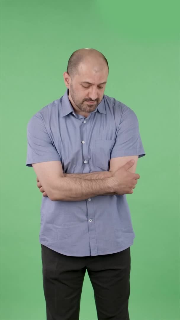 カメラを見ている中年男性の肖像画は、交差腕のジェスチャーを示しています。スタジオ内の緑のスクリーンにポーズをとった青いシャツに髭を生やした男。スローモーション。垂直ビデオ. — ストック動画