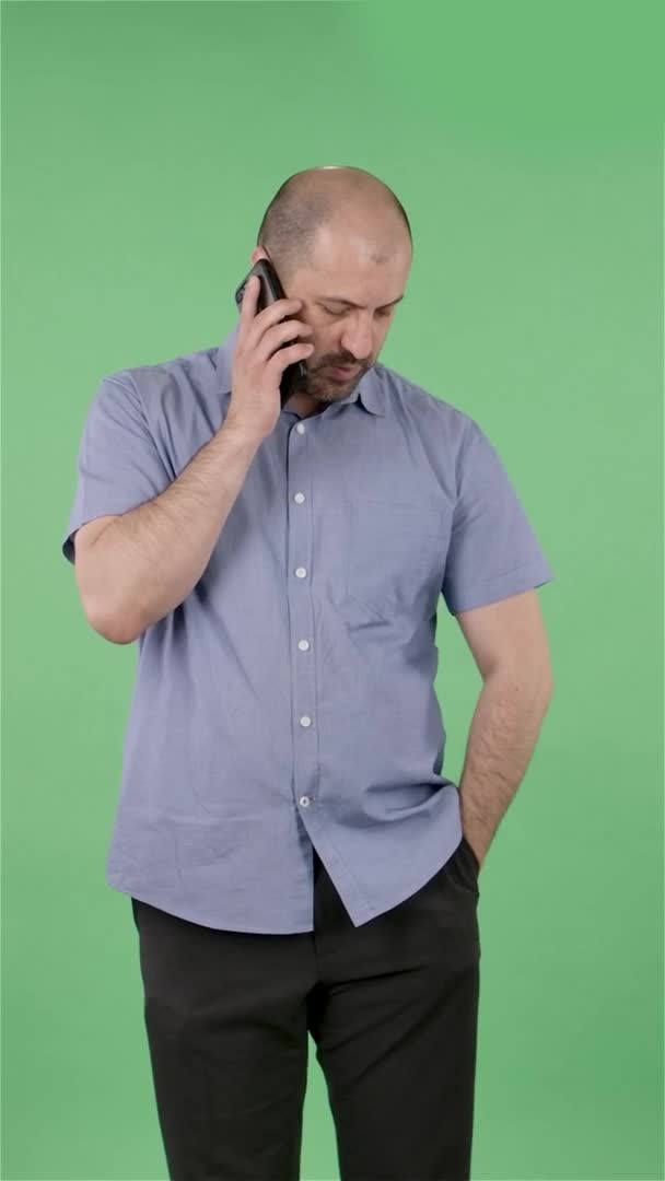 Retrato de un hombre de mediana edad hablando en su smartphone. Hombre calvo con barba en camisa azul posando en pantalla verde en el estudio. En cámara lenta. Vídeo vertical. — Vídeo de stock