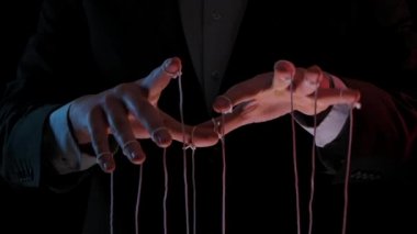Kuklacı parmaklarına bağlı iplerle kuklaları kontrol eder ve manipüle eder. Takım elbiseli iş adamı insanların zihinlerini ve hareketlerini kontrol eder. Erkek elleri siyah arkaplanda. Yavaş çekim.