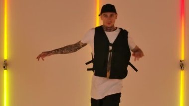 Dövmeleri olan genç bir adam, şık genç kıyafetleri ve şapkası olan, hip hop tarzında dans eden. Erkek dansçı stüdyodaki parlak neon ışıklarının arka planına karşı gösteri yapar. Kapatın. Yavaş çekim.