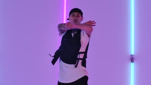 Junger Mann mit Tätowierungen, in stylischer Jugendkleidung und Mütze, der im Hip-Hop-Stil tanzt. Männliche Tänzer treten vor dem Hintergrund heller Neonlichter im Studio auf. Aus nächster Nähe. Zeitlupe. — Stockvideo