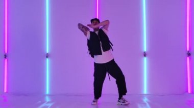 Dövmeleri olan genç bir adam, şık genç kıyafetleri ve şapkası olan, hip hop tarzında dans eden. Erkek dansçı stüdyodaki parlak neon ışıklarının arka planına karşı gösteri yapar. Yavaş çekim.