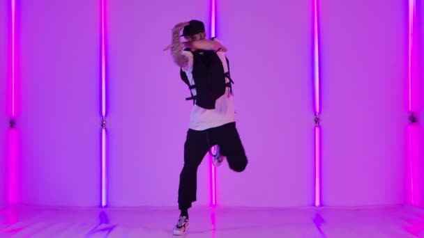 Stylový pouliční tanečník v mladistvých hipsterských šatech a čepice provádí komplexní hip hop taneční prvky. Mladý muž v tetováních cvičí své schopnosti na pozadí jasných neonových světel. Zpomalený pohyb. — Stock video