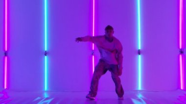 Dövmeli şık genç adam, parlak neon ışıkların önünde sokak dansı yapıyor. Hip hop tekniği öğretme konsepti. Gençlik alt kültürü. Çağdaş koreografi. Yavaş çekim.