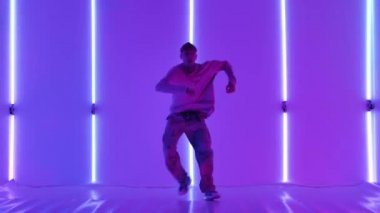 Dövmeli şık genç adam, parlak neon ışıkların önünde sokak dansı yapıyor. Hip hop tekniği öğretme konsepti. Gençlik alt kültürü. Çağdaş koreografi. Yavaş çekim.