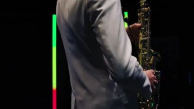Beyaz takım elbiseli bir saksafoncu karanlık bir stüdyoda neon lambaların arka planında gösteri yapıyor. Saksafoncu saksafoncu Jazzman. Bir müzisyenin gövdesi ve elleri yakın plan. Yörünge atışı. Yavaş çekim.