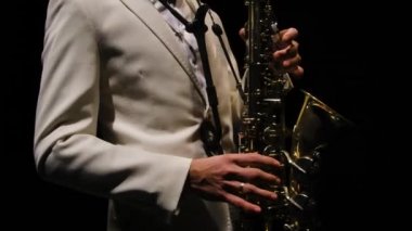 Beyaz takım elbiseli bir saksafoncu karanlıkta gösteri yapar. Saksafonlu Jazzman, arka planda siyah stüdyo arka planında. Bir müzisyenin gövdesi ve elleri yakın plan. Yörünge atışı. Yavaş çekim.