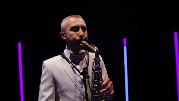 Kameran roterar runt snygg man i en vit kostym med fluga spelar saxofon. Saxofonist som uppträder i mörk studio mot bakgrund av neonlampor. Sakta i backarna. Närbild. — Stockvideo