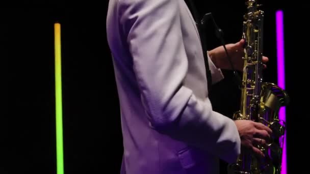 Saxophone player in a white suit performing against the backdrop of neon lamps in a dark studio. Saxophonist jazzman with sax. The torso and hands of a musician close up. Orbital shot. Slow motion. — Stock Video