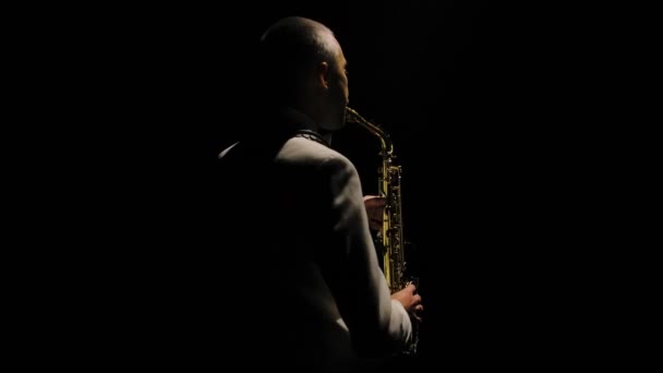 Caméra tourne autour de l'homme élégant dans un costume blanc joue le saxophone. Saxophoniste se produisant dans un studio sombre rétroéclairé. Au ralenti. Gros plan. — Video