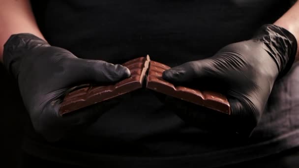 Chef chocolatero con guantes negros rompe la barra de chocolate por la mitad para derretir y hacer dulces, cocinando los postres dulces. Las manos de los hombres cierran. Movimiento lento. — Vídeos de Stock