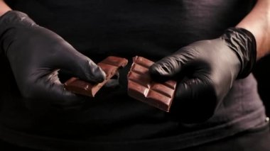 Siyah eldivenli adam çikolata kırıyor. Çikolatayı iki parçaya ayıran ellerinizi kapatın. Lezzetli çikolata, izole edilmiş siyah arka planda parçalara ayrılır. Yavaş çekim.