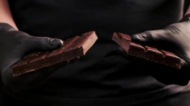 Siyah eldivenli adam çikolata kırıyor. Çikolatayı iki parçaya ayıran ellerinizi kapatın. Lezzetli çikolata, izole edilmiş siyah arka planda parçalara ayrılır. Yavaş çekim.