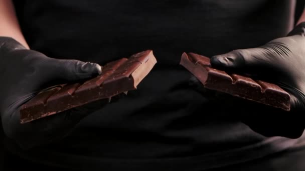 Hombre con guantes negros rompiendo chocolate. Cierre de manos rompiendo la barra de chocolate en dos partes. Delicioso chocolate se rompe en pedazos sobre fondo negro aislado. Movimiento lento. — Vídeo de stock