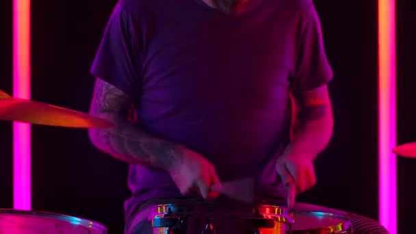 Skilled man muzikant speelt hedendaagse drumstel in een donkere studio verlicht door veelkleurige neon buizen. Close-up van een drummers getatoeëerde handen slaan drums met houten stokken. Langzame beweging. — Stockvideo