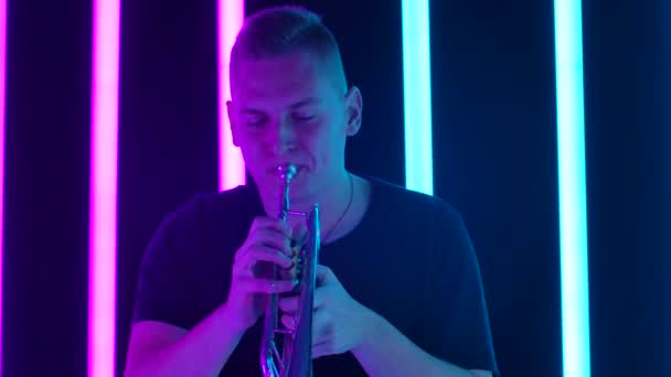 Um homem toca trompete no estúdio contra um fundo de tubos de néon rosa e azul. Desempenho clássico no tubo. Movimento lento. Fechar. — Vídeo de Stock