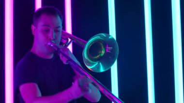 Tromboncu müzik çalar, hava üfler ve tüp hareket eder. Stüdyoda trombon çalan bir adam mavi ve pembe neon tüplerin arka planına karşı. Kapatın. Yavaş çekim.