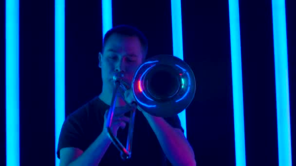 Desempenho solo de um músico tocando um trombone de bronze clássico. Um homem se apresenta em um estúdio escuro contra o pano de fundo de lâmpadas de néon multicoloridas. Fecha. Movimento lento. — Vídeo de Stock