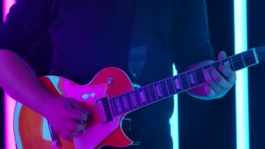 Bir müzisyen, turuncu bir elektro gitarla stüdyodaki mavi ve pembe neon boruların arka planına karşı solo çalıyor. Bir erkeğin elleri, telleri koparıp boynuna dokunurken. Yavaş çekim.