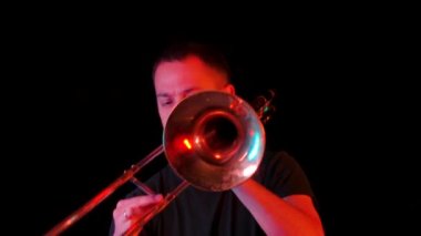 Stüdyoda trombon çalan bir müzisyenin ön görüntüsü neon ışıkta siyah bir arka plana karşı. Trombist, bir rüzgar enstrümanında ska punk tarzında müzik çalıyor. Kapatın. Yavaş çekim.
