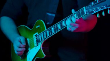 Bir grup gitarist, enstrümanlarıyla stüdyoda neon ışıkta siyah bir arkaplanda beste yapıyor. İpleri koparan müzisyenlerin ellerini kapatın. Yavaş çekim.