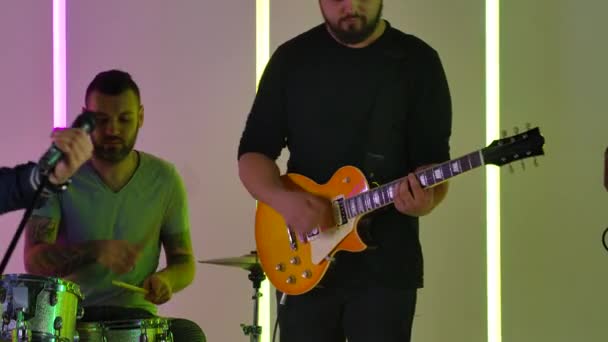 La banda interpreta su música en el estudio con el telón de fondo de luces de neón multicolores. El guitarrista y el baterista tocan sus instrumentos profesionalmente. En cámara lenta. De cerca.. — Vídeos de Stock