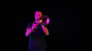 Stüdyoda trombon çalan bir müzisyenin ön görüntüsü neon ışıkta siyah bir arka plana karşı. Trombist, bir rüzgar enstrümanında ska punk tarzında müzik çalıyor. Kapatın. Yavaş çekim.