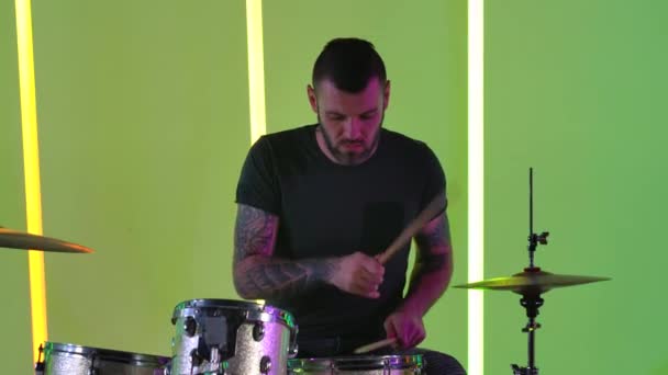 Expressieve mannelijke muzikant speelt drums in de studio tegen een achtergrond van veelkleurige neon buizen. Een drummer met tatoeages op zijn armen speelt rockmuziek. Muziekconcept. Langzame beweging. Sluiten.. — Stockvideo