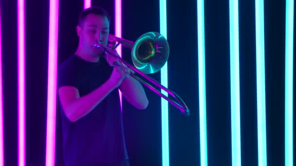 Тромбоніст грає музику, дме повітря, а трубка рухається. Чоловік, який виконує соло на тромбоні в студії на тлі синіх і рожевих неонових трубок. Близько. Повільний рух . — стокове відео