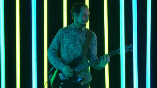 O artista toca uma guitarra elétrica preta e canta uma música em um estúdio escuro cercado por lâmpadas de néon multicoloridas brilhantes. Uma pessoa contente que gosta de rock. Fecha. Movimento lento. — Vídeo de Stock