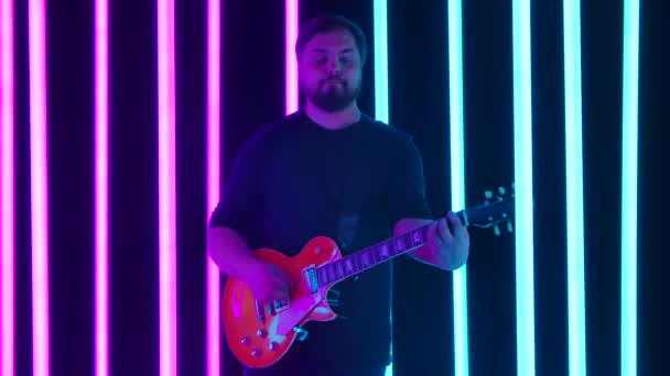 Gitarristen njuter av sitt elgitarrspel i en mörk studio omgiven av klarblå och rosa neonrör. En man lär sig nya ackord på sitt instrument. Sakta i backarna. Närbild. — Stockvideo