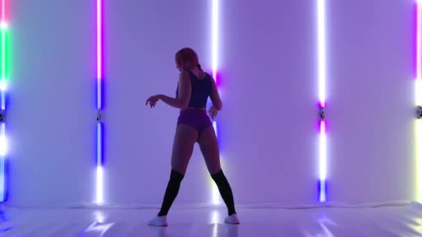 Sexig ung kvinna dansar twerk. Sensuell kvinna i korta shorts flyttar sin kropp och twerkar mot bakgrund av ljusa neonljus i studion. Långsamma rörelser. — Stockvideo