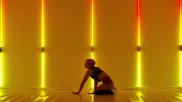 Beautiful sexy woman dancing twerk sitting on floor. Dancer shakes her ass, dances twerk. Sensual female dancing booty in studio against backdrop of bright yellow and red neon lights. Close up. Slow — Stock video