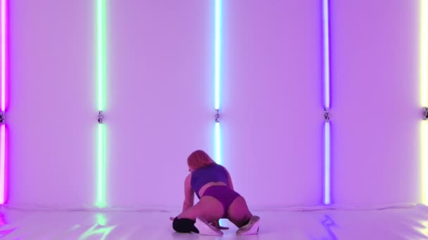 Sensual athletic woman shaking her ass, dancing twerk in the studio. Sexy dancer twerks in short shorts, sitting and moving her booty against the backdrop of bright neon lights. Slow motion. — Vídeo de stock