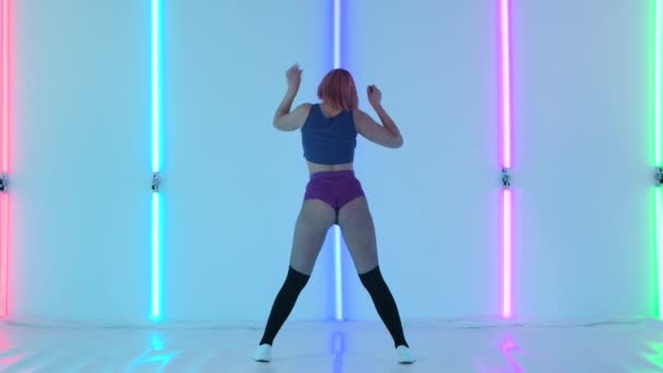 Jovem mulher com cabelo rosa com um corpo sexy dança twerks suas nádegas. Dança erótica de uma mulher sensual contra o fundo de luzes de néon brilhantes no estúdio. Fecha. Movimento lento. — Vídeo de Stock