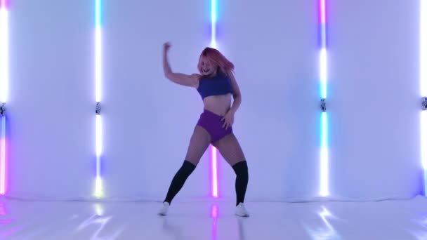 Sexig ung kvinna dansar go-go eller twerk. En smal kvinnlig dansare utför en erotisk dans, förföriskt vrider på rumpan. Booty dans i studion med ljusa neonljus. Långsamma rörelser. — Stockvideo