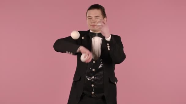 黒と白のスーツの若い男は3つのボールでジャグリングします。サーカスのパフォーマーはピンクの背景のスタジオで集中力、スキル、反応を練習します。閉じ込められた。スローモーション. — ストック動画