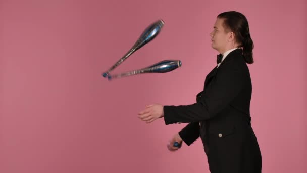 Sidovy av professionell cirkusartist jonglerar mästerligt med klubbar i studion på en rosa bakgrund. Man i svart och vit kostym övar koncentration, skicklighet och skicklighet. Närbild. Långsamma rörelser. — Stockvideo